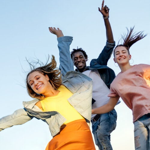 Drei junge Erwachsene springen fröhlich in die Luft, mit einem breiten Lächeln, erhobenen Armen und dem Wind in den Haaren vor einem klaren blauen Himmel. Sie wirken glücklich und voller Energie und genießen einen unbeschwerten Moment zusammen.
